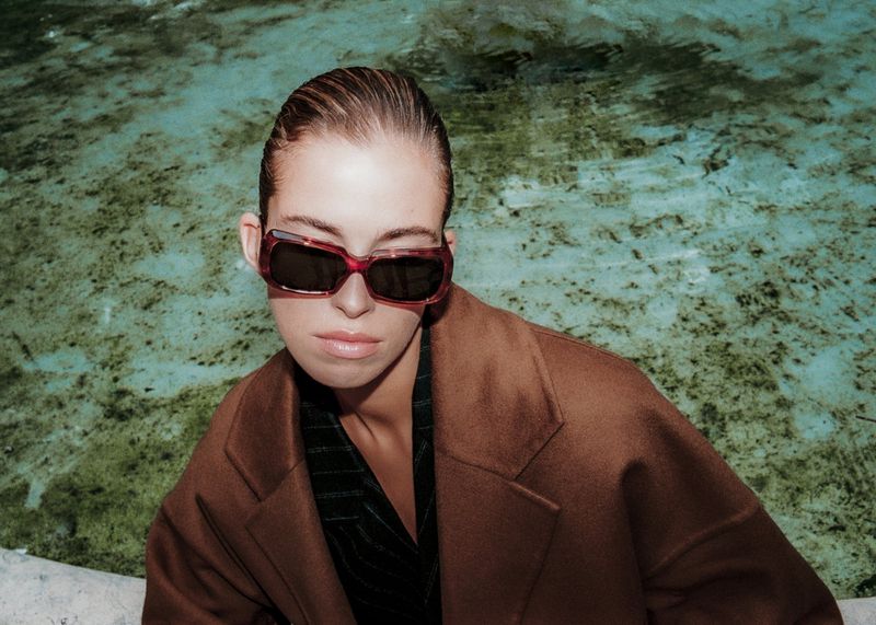 A stylish woman in sunglasses and a brown coat sits in front of a textured, light-blue stone fountain with slicked-back hair.