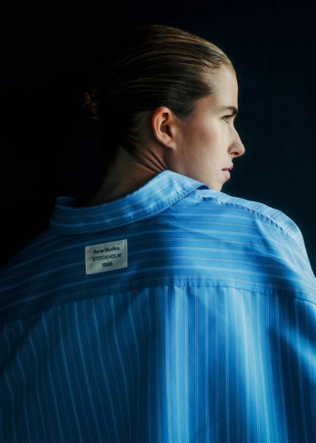 Woman in a blue-striped shirt with an Acne Studios label on the back, side profile with light hair against a dark background.