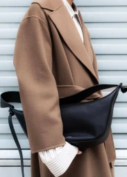 Cropped view of a model in a camel wool coat carrying a black leather crossbody bag against a pale slatted background.