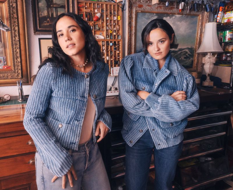 Two women wearing matching blue pinstripe jackets and jeans pose indoors in front of vintage shelves and framed artwork.