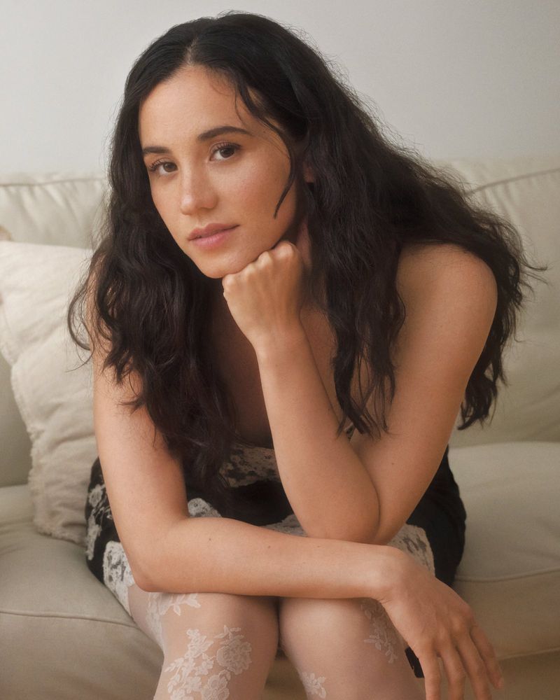 Portrait of a young adult woman with long dark hair sitting indoors, looking at the camera with hand on chin, wearing a black dress.