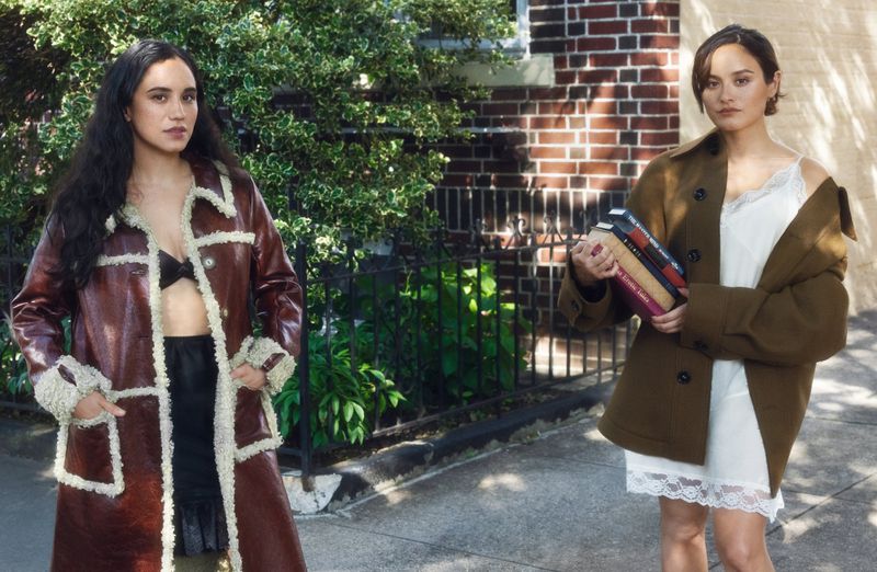 Two adult women standing outdoors next to a brick building, both wearing fashionable outerwear, with a book in one woman's arms.