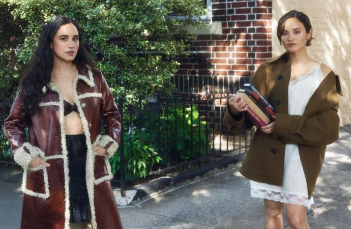 Two adult women standing outdoors next to a brick building, both wearing fashionable outerwear, with a book in one woman's arms.