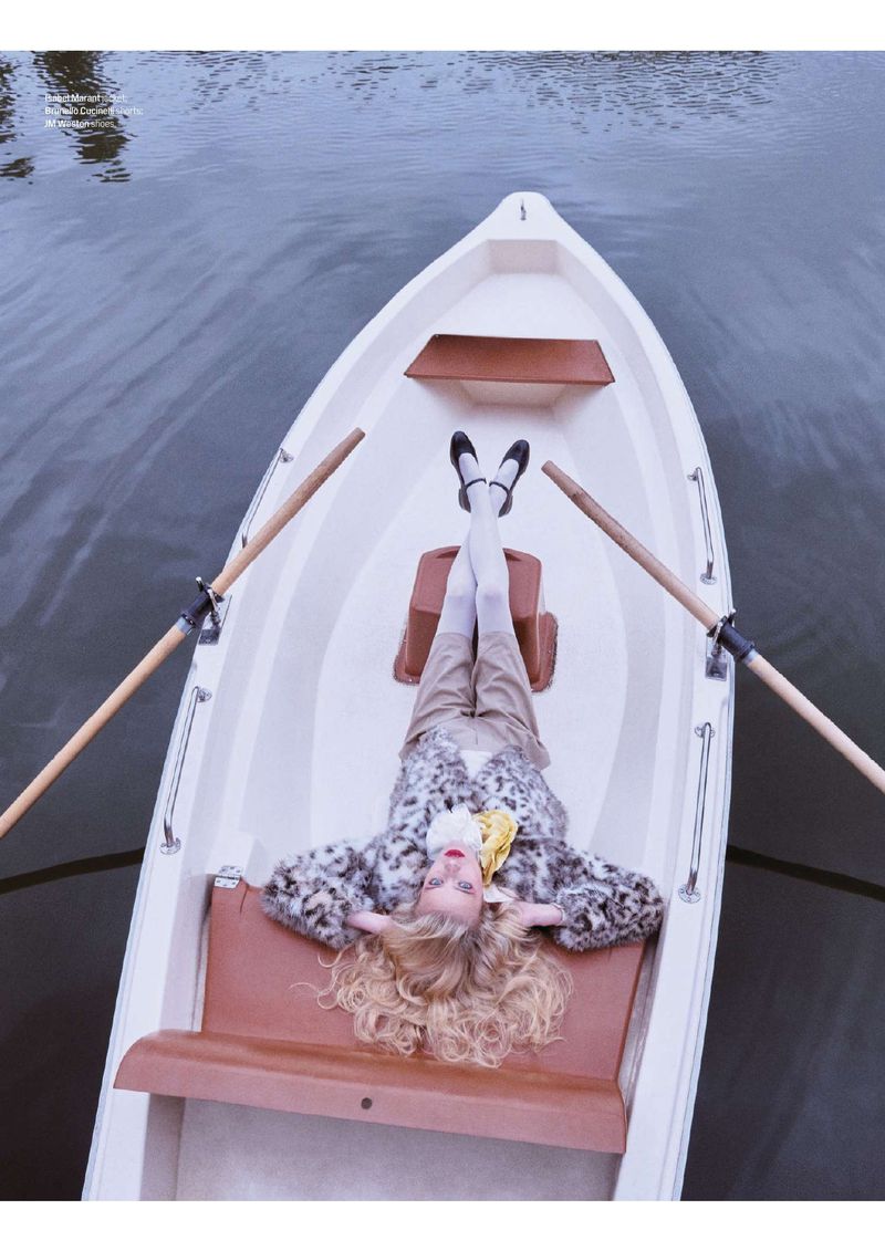 A young woman with blonde hair lounges in a rowboat on the water. She wears a stylish outfit with flats and a patterned coat.