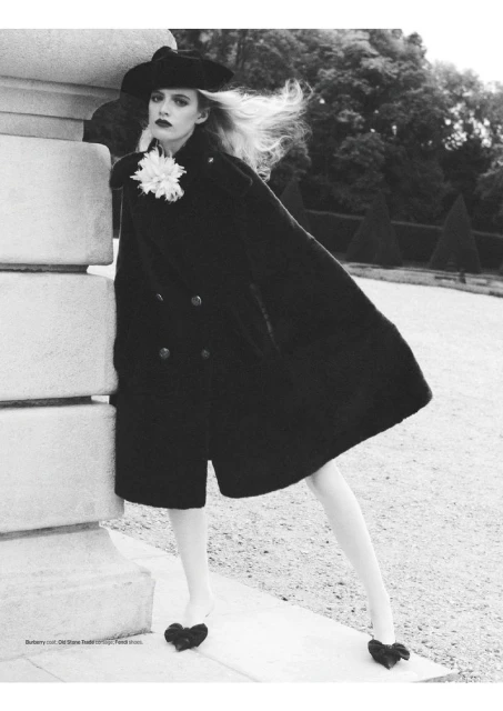 A woman in a black coat and hat stands next to a stone column in a monochromatic outdoor fashion shot with trees in the background.