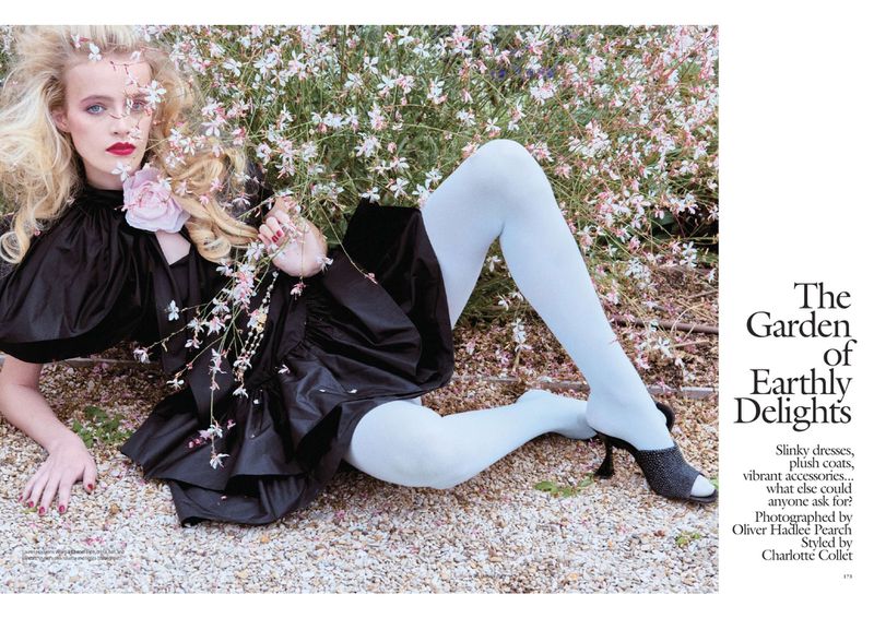 Blonde woman in black dress lying amongst white flowers in a fashion editorial. She wears white tights and black heels.