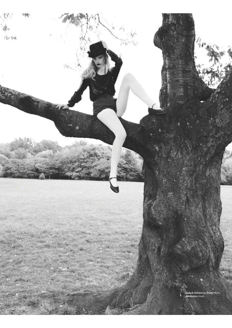 A woman in a black hat and shorts sits on a tree branch in a grayscale shot with a grassy background and light sky.