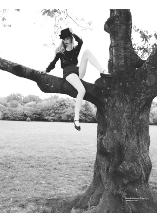 A woman in a black hat and shorts sits on a tree branch in a grayscale shot with a grassy background and light sky.