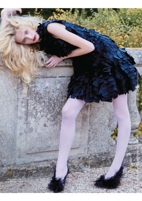 Fashion editorial shot of a blonde woman in a black dress with feathered heels, leaning on a stone wall in an outdoor setting. 