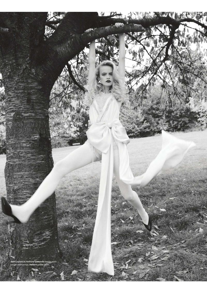 Monochrome shot of a blonde woman hanging from a tree branch in a lingerie dress, stockings and heels in an outdoor setting with grass.