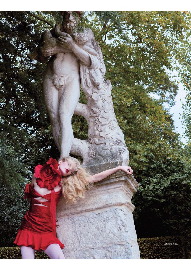 Woman in a red dress poses next to a stone statue in an outdoor park-like environment, featuring greenery and classical art.