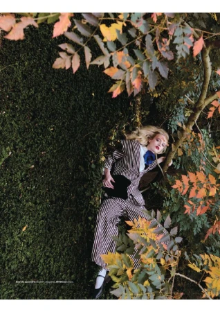 Adult woman in a striped suit poses in a forest environment with trees, greenery, and orange leaves in an editorial fashion setting.