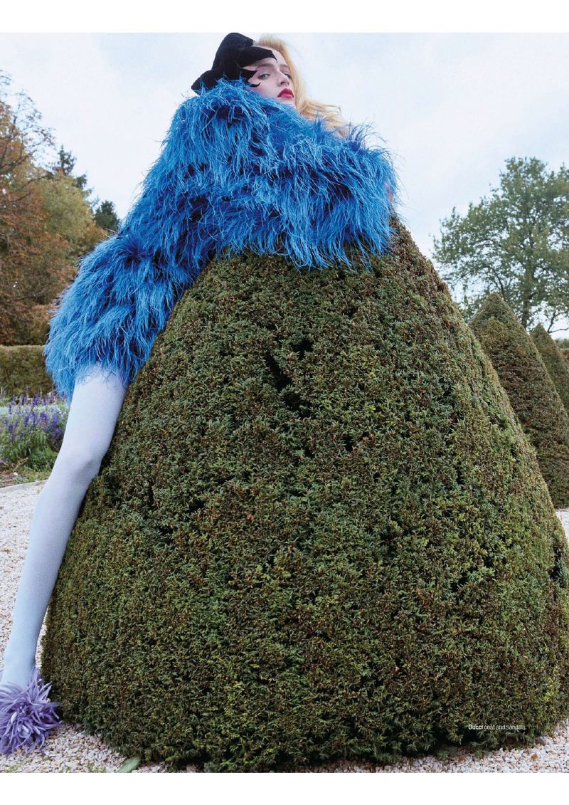 Fashionable woman in a blue feathered coat poses outdoors in a garden with sculpted bushes, wearing white tights and purple feathered shoes.