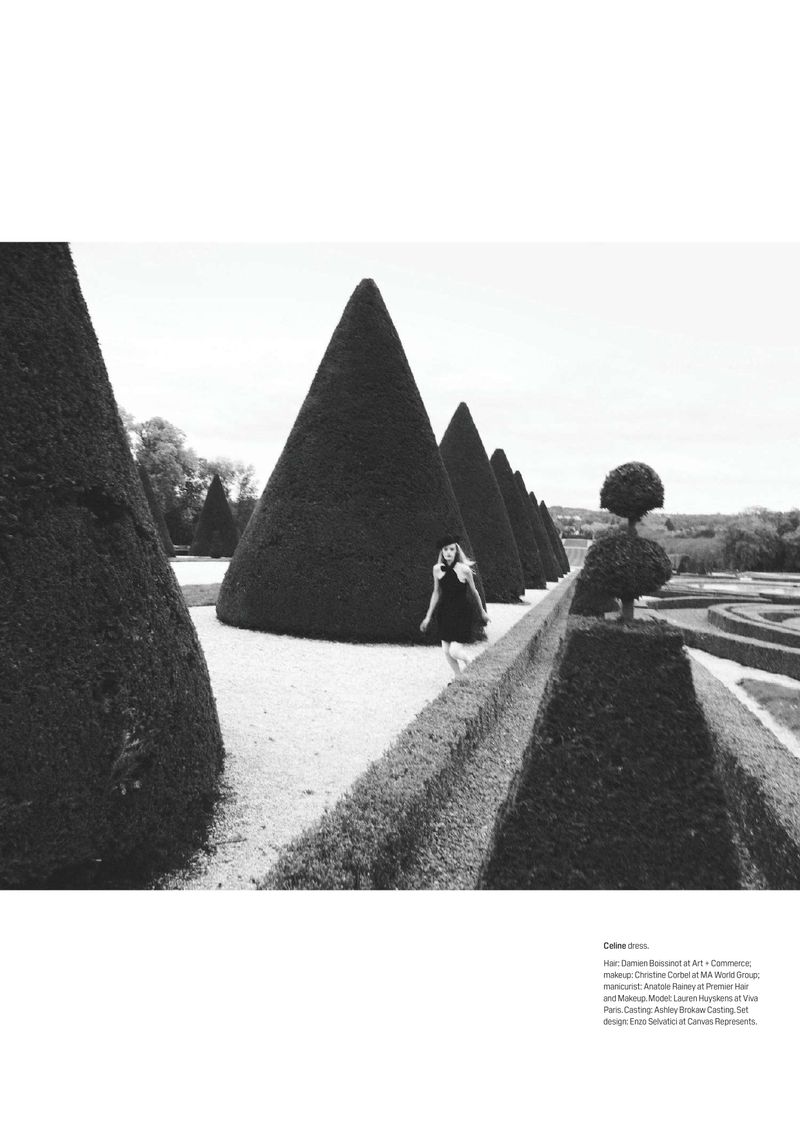 Black and white outdoor fashion shot of a woman in a dress walking in a formal garden with trimmed conical trees in a row.