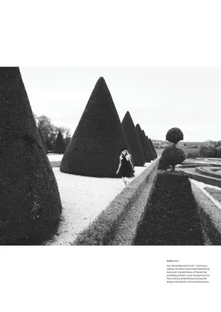Black and white outdoor fashion shot of a woman in a dress walking in a formal garden with trimmed conical trees in a row.