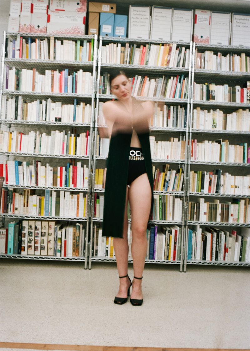 A woman in a black dress and heels strikes a blurry pose in front of tall bookshelves filled with books in an indoor setting.