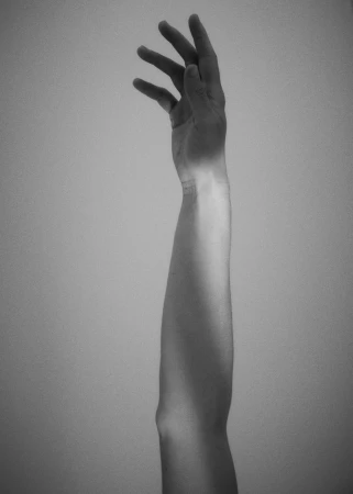Black and white photo of a raised adult arm, fingers extended, showcasing muscle and bone structure in a minimalist studio setting.