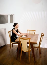 Woman in a black dress seated at a wooden dining table with a single egg on a plate in an interior room