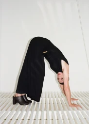 Model in black jumpsuit bending backwards into a bridge pose wearing dark mules against a white studio background