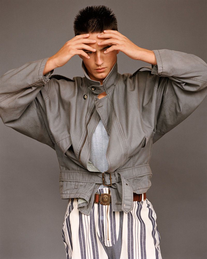 Young adult in gray jacket and striped pants with hands on head, posing in a studio setting against a neutral gray background.