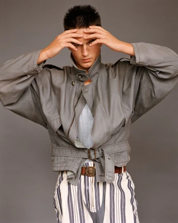 Young adult in gray jacket and striped pants with hands on head, posing in a studio setting against a neutral gray background.