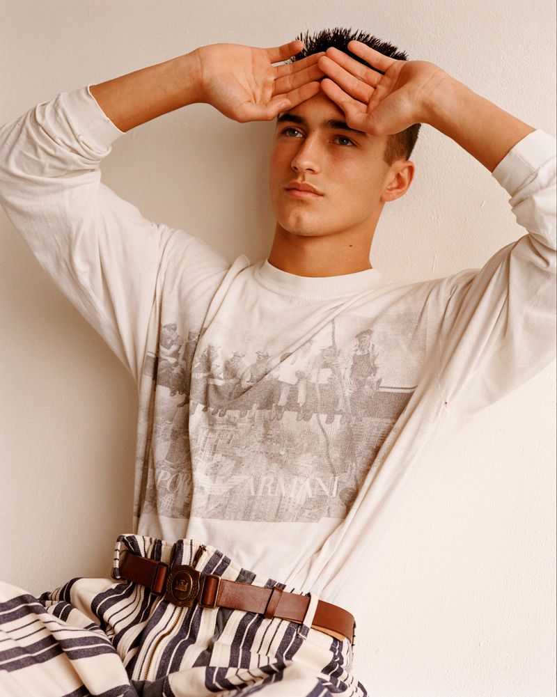 Young man poses indoors with hands framing face. He wears a printed long-sleeve shirt, striped pants, and a brown belt.