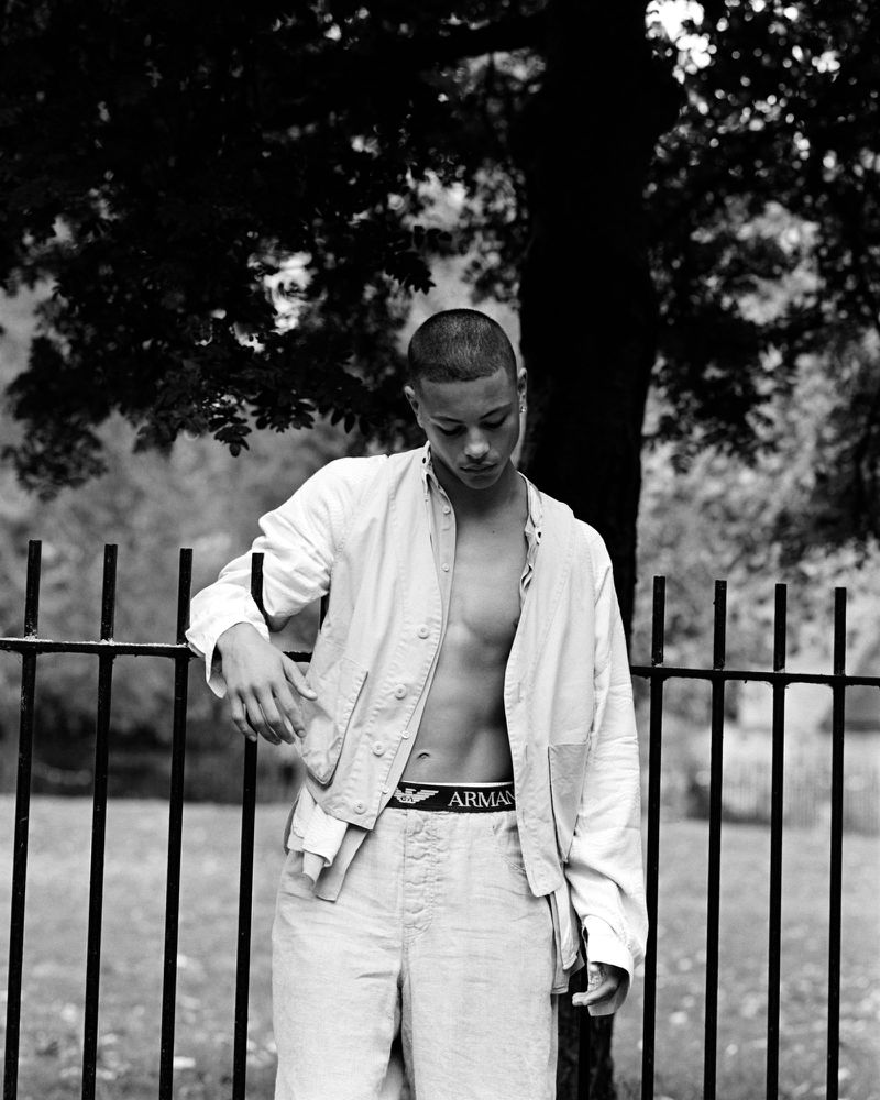 Young man poses leaning on a black metal fence, wearing a white shirt and linen pants, showcasing his torso in a black-and-white outdoor shot.