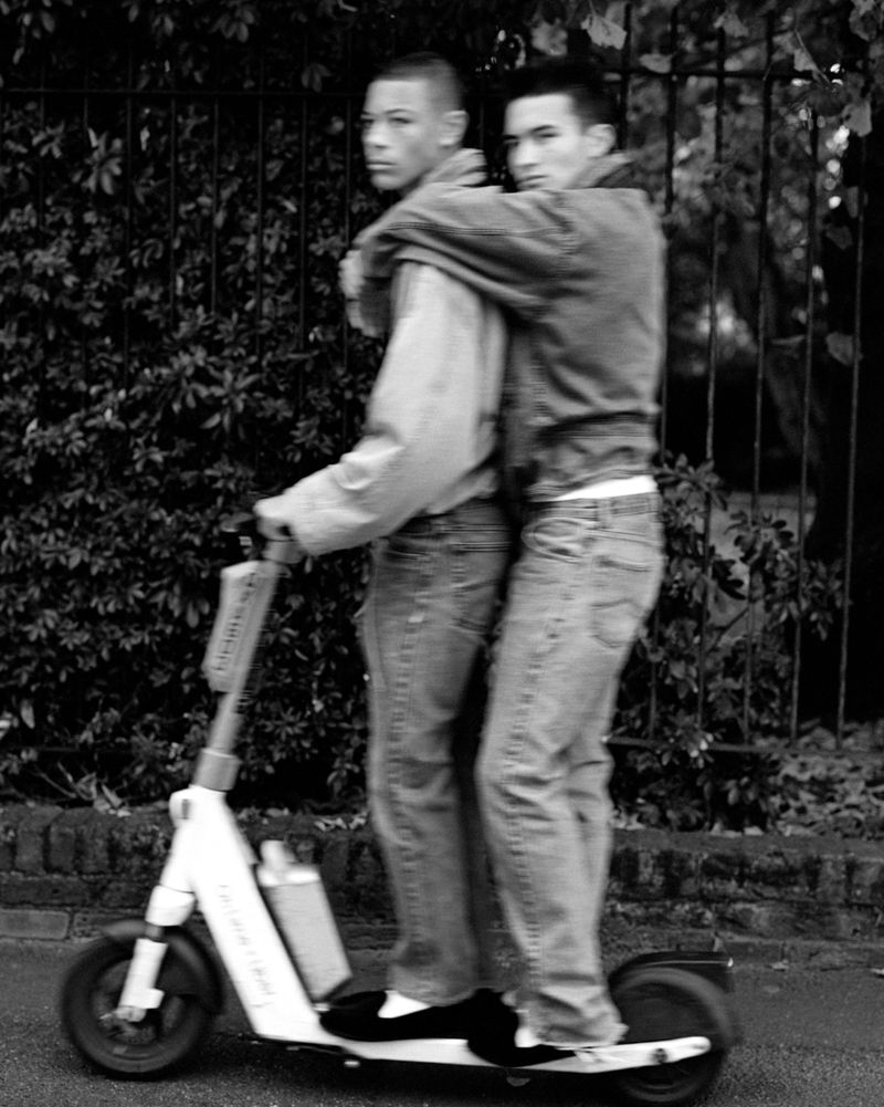 Two young adult men share an electric scooter outdoors on a paved path, one standing behind the other, in a black and white image.