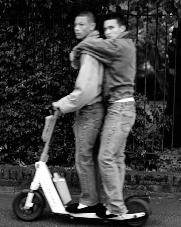 Two young adult men share an electric scooter outdoors on a paved path, one standing behind the other, in a black and white image.