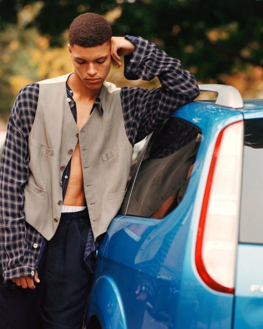 A young adult leans casually against the back of a blue car, wearing an unbuttoned vest and a plaid shirt against a natural outdoor setting.