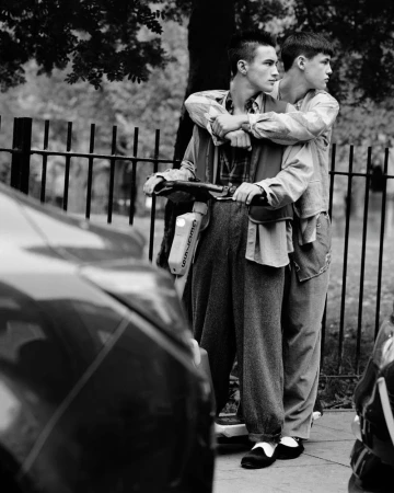 Two young men stand side-by-side outdoors, one with his arm around the other, near a road with cars, in a black and white photograph.