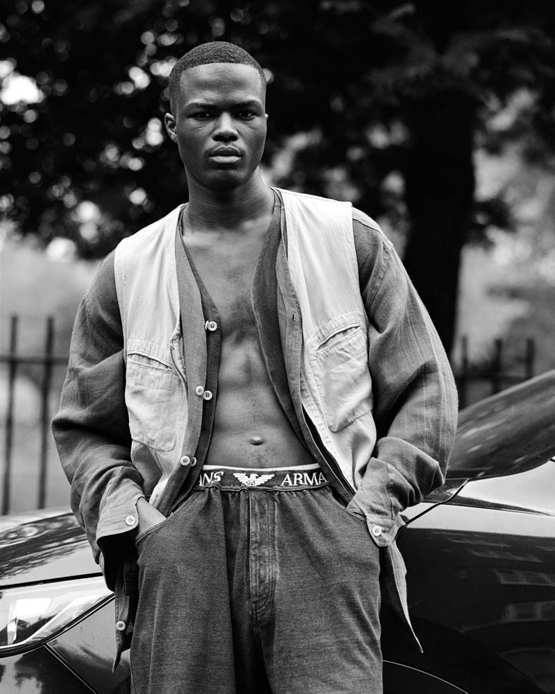 Monochrome portrait of a Black man in a vest and trousers, posing in front of a dark-colored car with trees in the background.