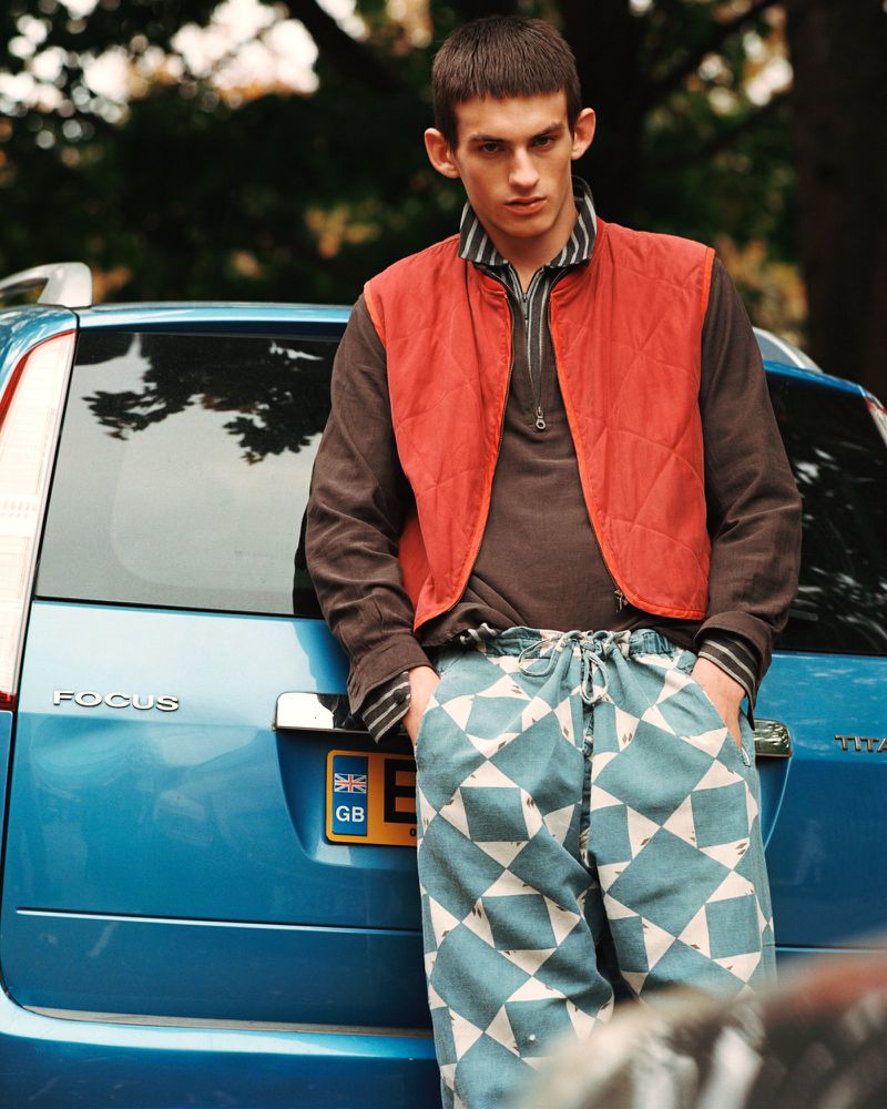 A young adult male model stands in front of a light blue car wearing a burnt orange vest and patterned pants in a fashion editorial.