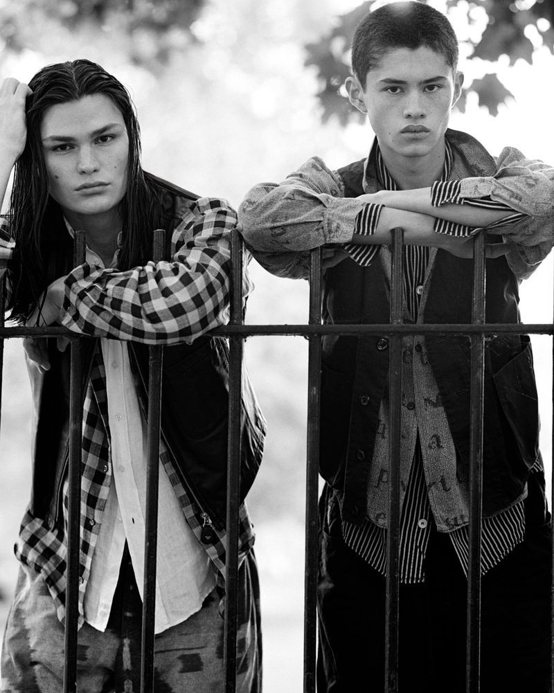 Two young men stand side-by-side in a black and white medium shot, leaning against a black iron fence outdoors with trees in the background.