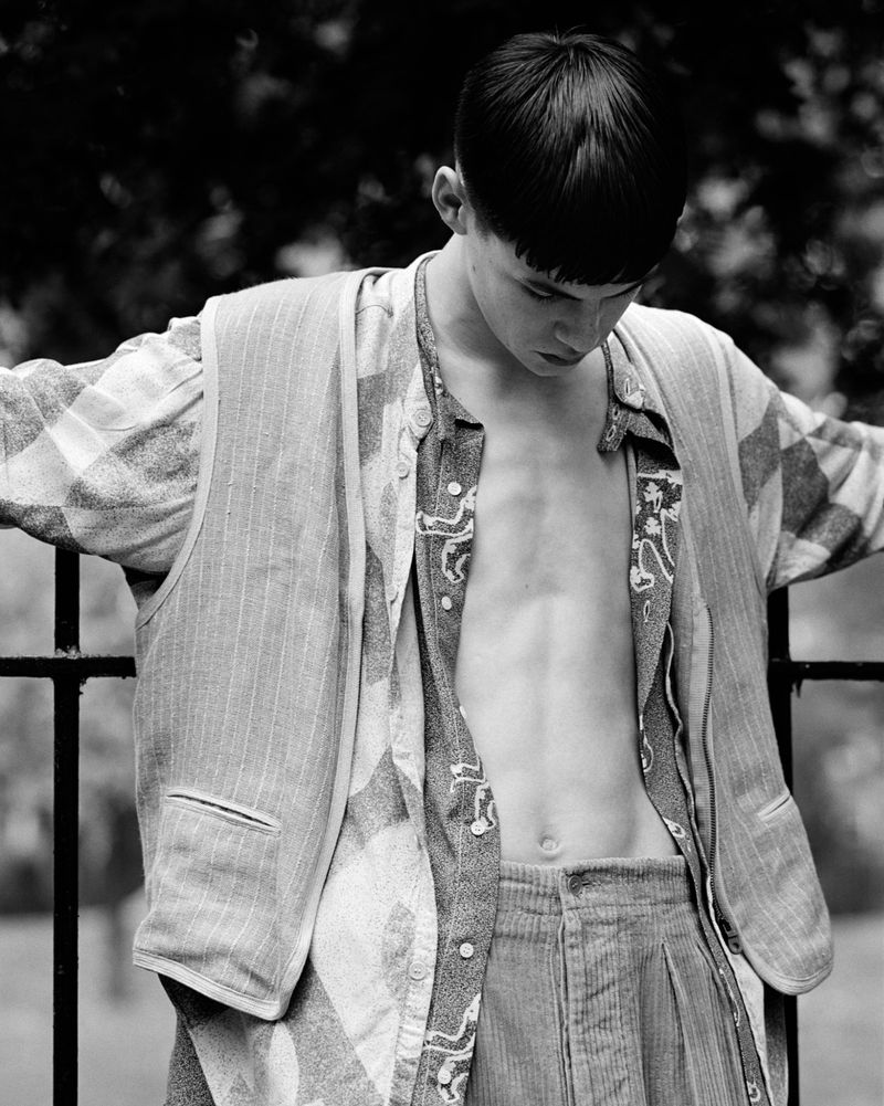 Black-and-white portrait of a young adult male in an open printed shirt, vest, and corduroy pants, posed outdoors against foliage and metal railing.