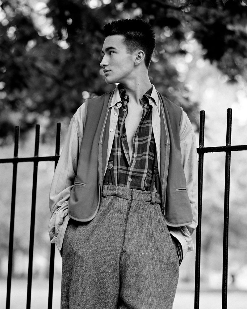 A young man poses outdoors wearing a buttoned plaid shirt, vest and wide-legged tweed pants, standing near a black metal fence.