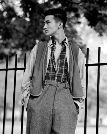 A young man poses outdoors wearing a buttoned plaid shirt, vest and wide-legged tweed pants, standing near a black metal fence.