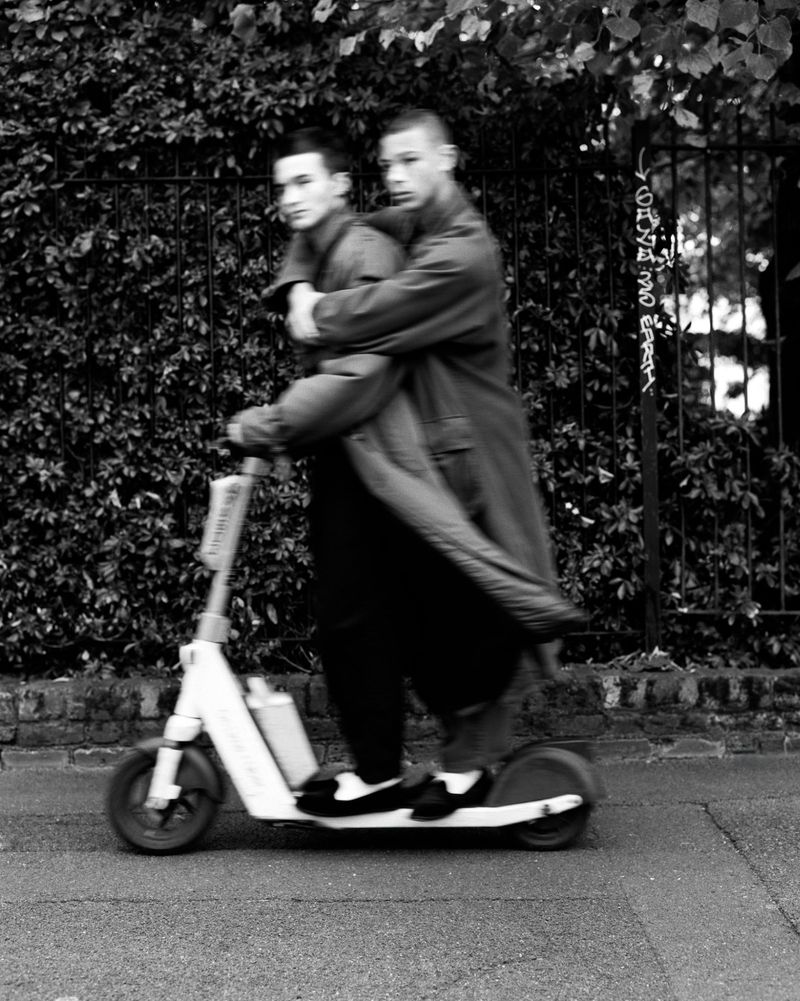 Two young adult men riding an electric scooter in a monochrome street scene with motion blur, showcasing an urban lifestyle moment.