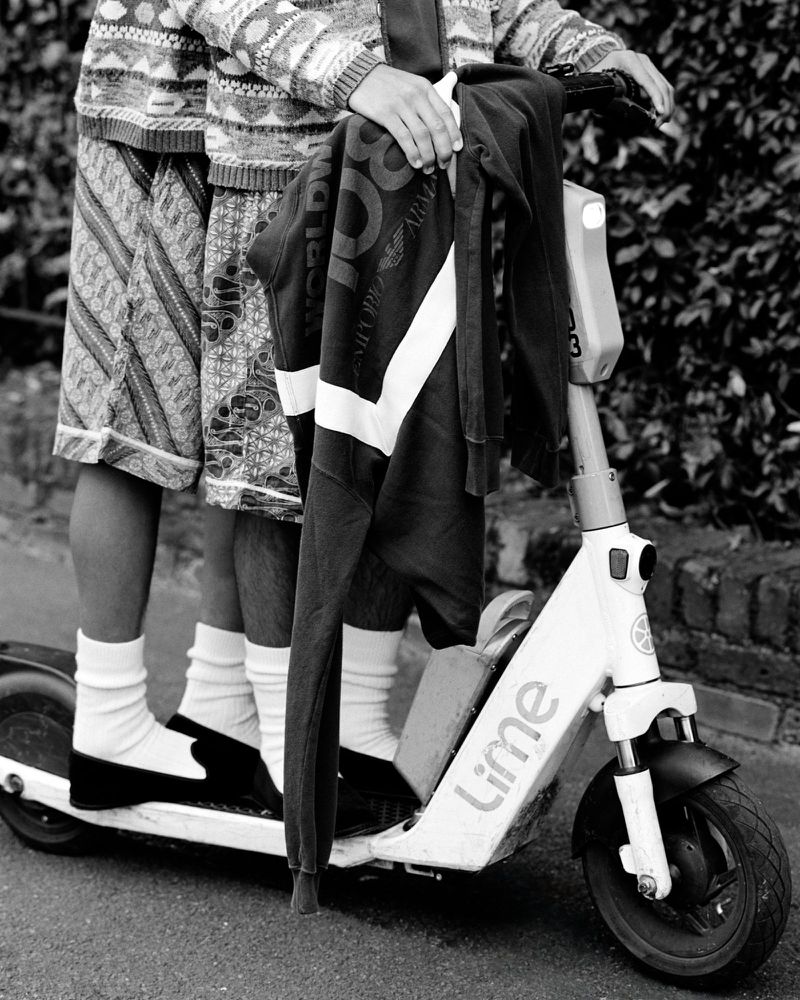Two people ride a Lime electric scooter on a street, one wearing a patterned sweater and shorts, the other holding a black jacket.