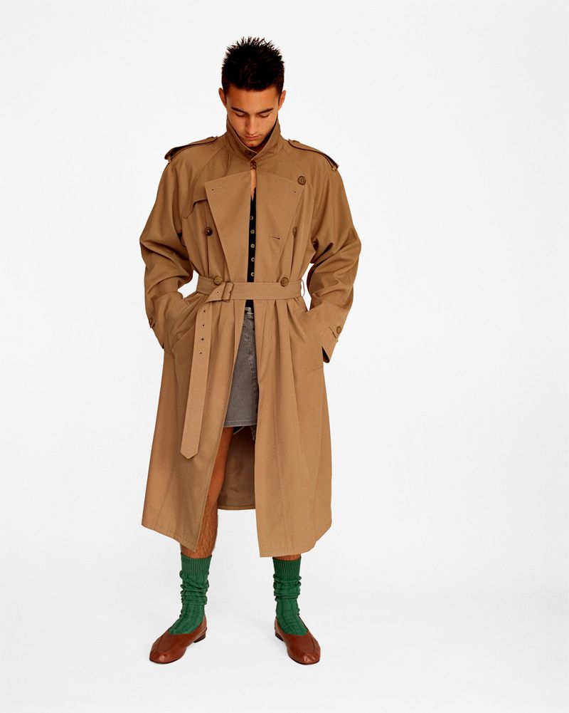 Man in trench coat, visible gray shorts, and green socks stands in a white studio. Full shot with casual pose and neutral expression.