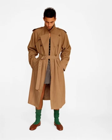 Man in trench coat, visible gray shorts, and green socks stands in a white studio. Full shot with casual pose and neutral expression.