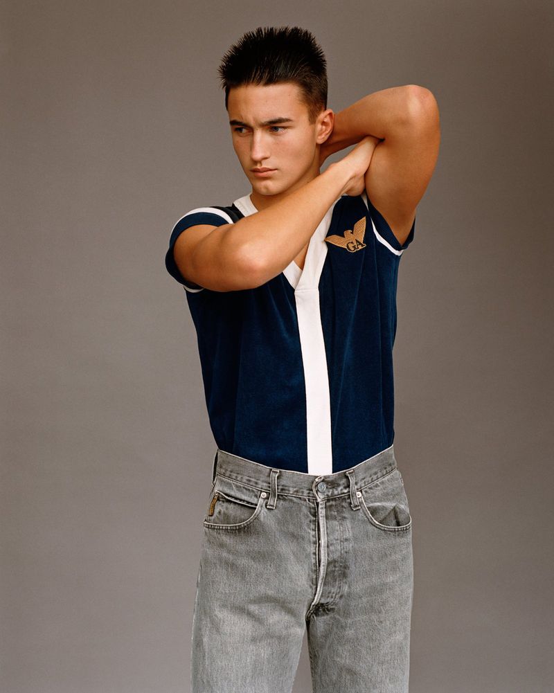 Portrait of a handsome adult male with hands behind his head wearing a navy-and-white shirt and grey jeans against a grey backdrop.