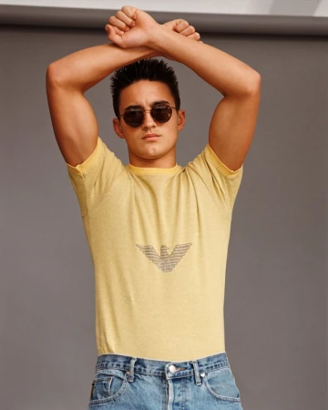 Portrait of a young adult man with arms raised wearing a yellow tee shirt, jeans, and sunglasses in a studio. Fashion editorial.
