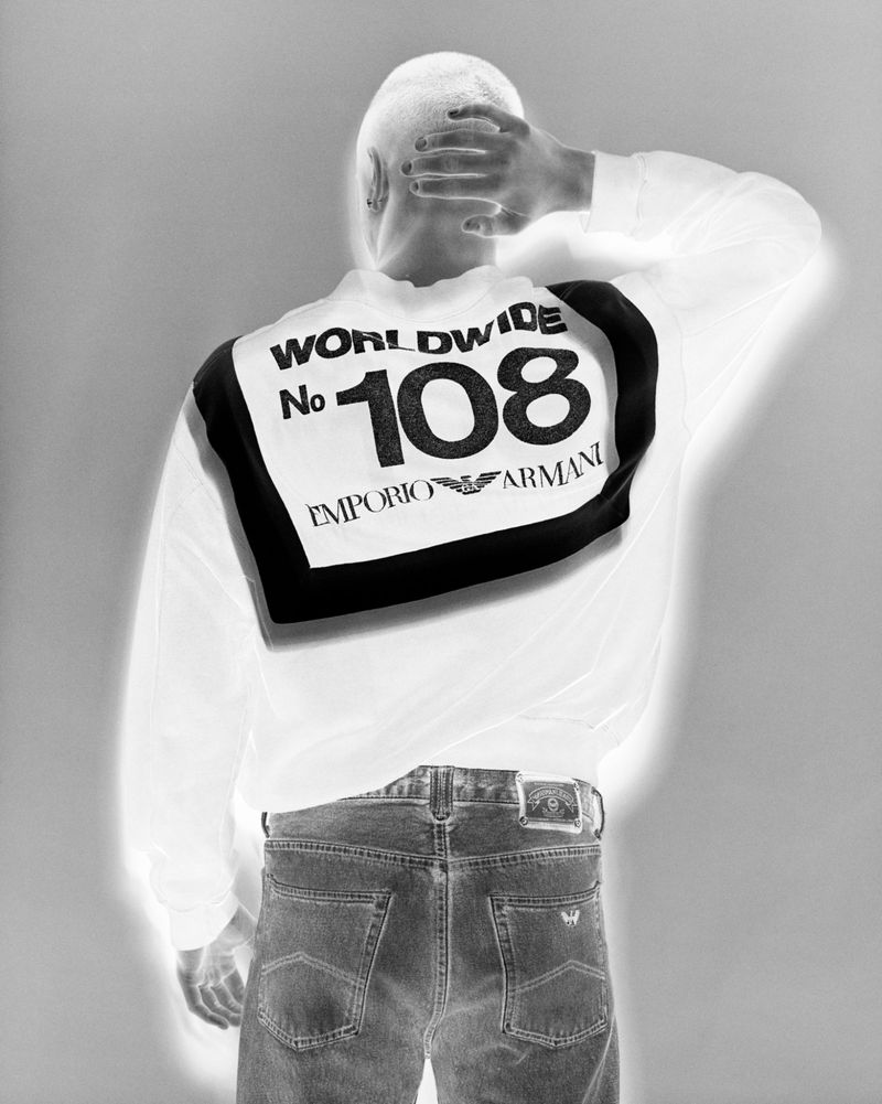 An adult leans forward, showcasing a white Emporio Armani long-sleeved shirt and dark-wash jeans, set against a grayscale backdrop.