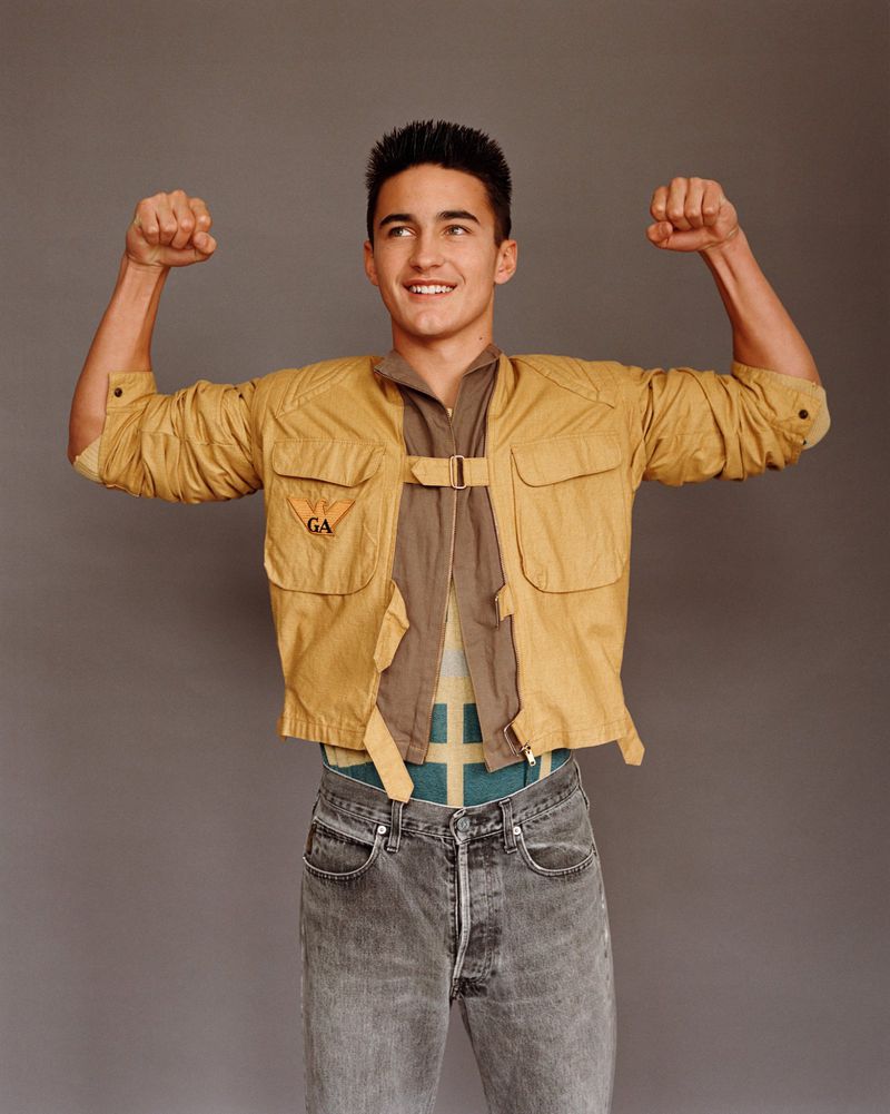 A young adult man with dark hair and a bright smile flexes his arms, wearing a tan jacket, patterned top, and gray jeans against a gray backdrop.
