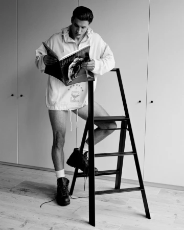 Monochrome portrait of a man reading a magazine while standing on a black stepladder in a minimalist room with white walls and wood floors.