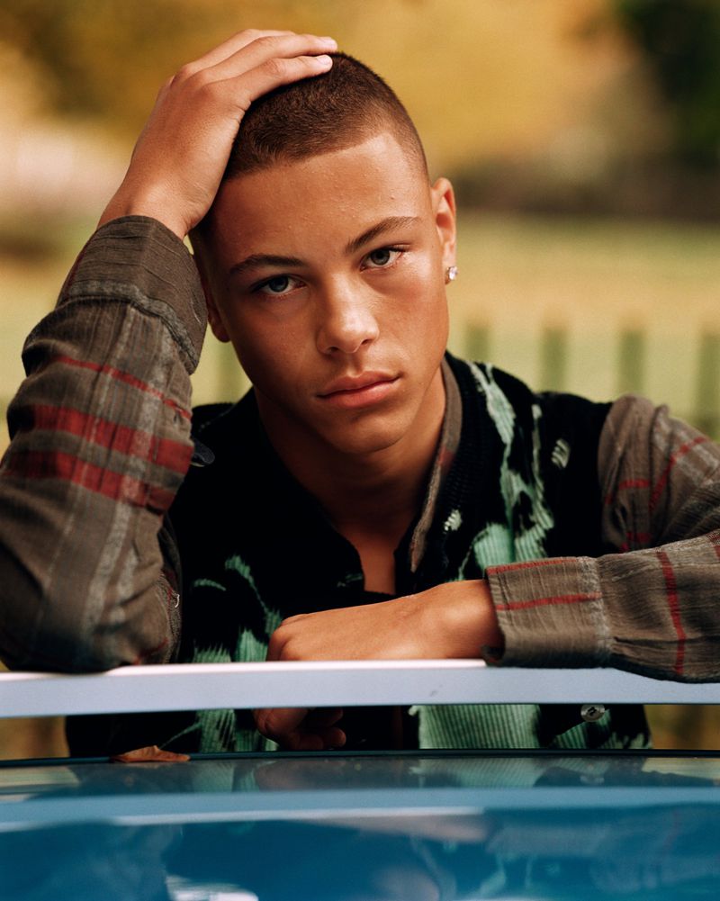 Portrait of a young adult male with short hair, wearing a patterned shirt and vest, with his hand on his head.