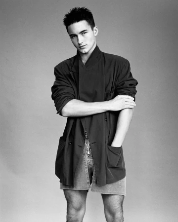Monochrome portrait of a male model wearing an oversized dark jacket and denim shorts against a plain grey studio backdrop.