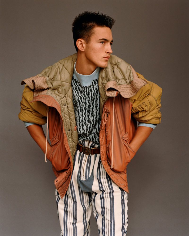 Young adult male fashion model poses in striped pants, colorful puffer jacket in a studio portrait with a neutral gray backdrop.