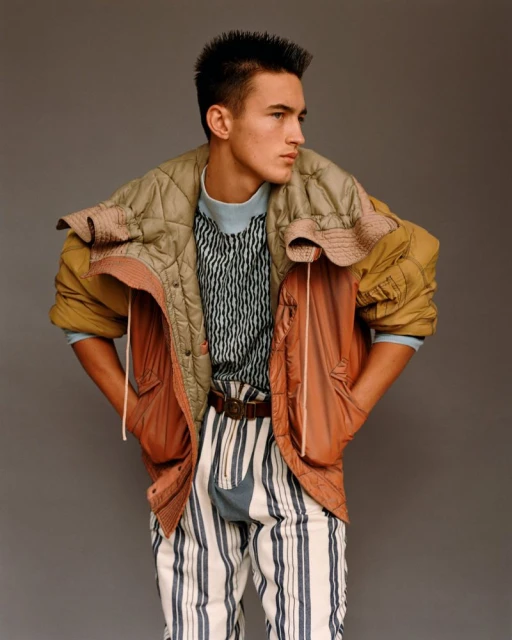 Young adult male fashion model poses in striped pants, colorful puffer jacket in a studio portrait with a neutral gray backdrop.